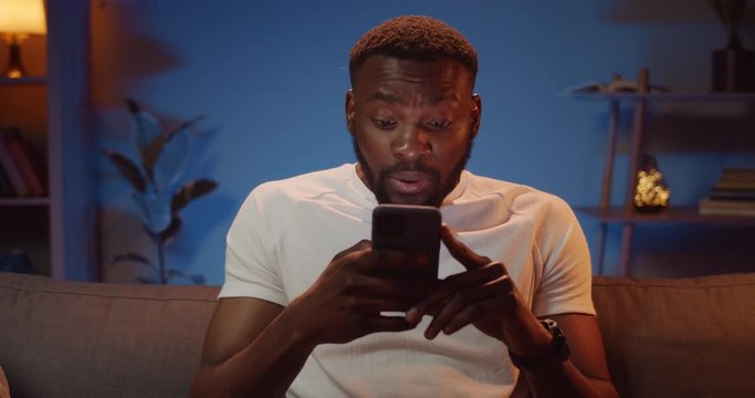 Handsome young guy using his smartphone and looking happy when getting good news. Bearded african man holding mobile phone in hands and making surprised face while sitting on sofa.