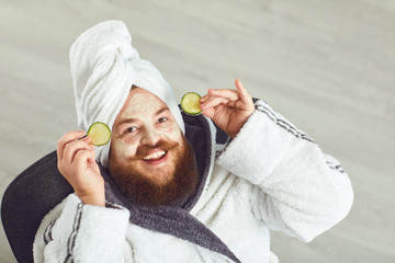 Funny fat bearded man with a cosmetic mask on his face in bathrobe towel on his head on his face resting against a gray background - obrazy, fototapety, plakaty