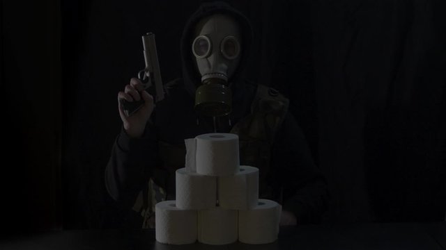 Gasmask wearing Man behind stack of toiletpaper rolls cocking gun and aiming it towards camera
