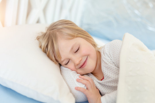 Cute adorable caucasian blond little toddler girl lying in bed on white pillow. Portrait of small beautiful long haired happy baby sweet dreaming asleep and smiling at bedtime in room at home