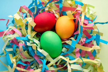 Three dyed Easter eggs in a colorful nest on mixed blue and yellow background. Top view. Symbol of main Christian feast celebration. Easter handcraft with colored paper.