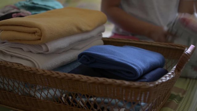 Woman is folding clothes and putting into a wooden basket. Housewife doing some house work in bedroom. Close up a pile of folded clothes on the bed.