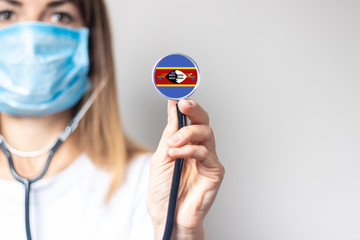 female doctor in a medical mask holds a stethoscope on a light background. Added flag of Eswatini....