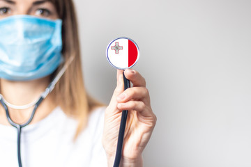 female doctor in a medical mask holds a stethoscope on a light background. Added flag of Malta....