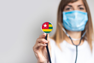 female doctor in a medical mask holds a stethoscope on a light background. Added flag of Togo. Concept medicine, level of medicine, virus, epidemic