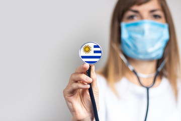 female doctor in a medical mask holds a stethoscope on a light background. Added flag of Uruguay....