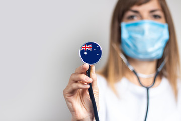 female doctor in a medical mask holds a stethoscope on a light background. Added flag of Australia....