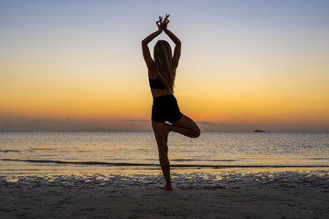 Fototapeta na wymiar Silhouette of woman standing at yoga pose on the tropical beach during sunset. Girl practicing yoga near sea water