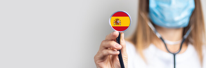 female doctor in a medical mask holds a stethoscope on a light background. Added flag of Spain....
