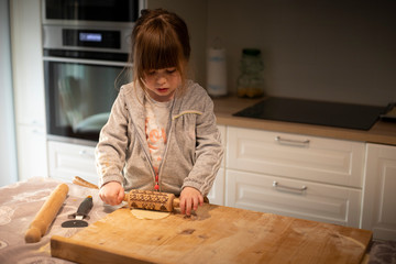 Child girl in white kitchen flattening pizza dough with a small rolling pin on a wooden board