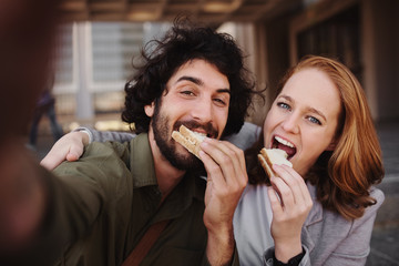 Cheerful young professional couple taking a selfie using smartphone while grabbing a bite of sandwich