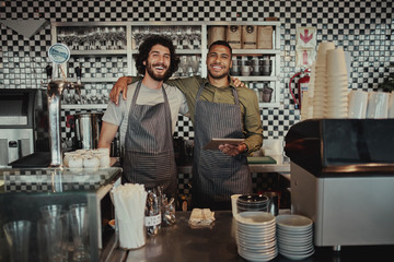 Portrait of smiling colleagues standing behind counter in cafe holding digital tablet with hands on each other shoulder looking at camera