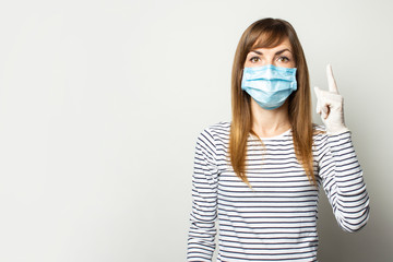 Young woman in protective medical mask and latex gloves points her finger up on a light isolated...