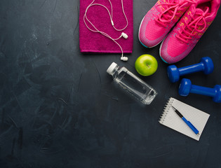 Fitness concept with sneakers headphones dumbbells bottle of water and apple on black concrete background