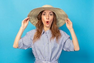 surprised young girl in a dress and hat, holds a hat with hands on a blue background. Concept of vacation, summer and travel