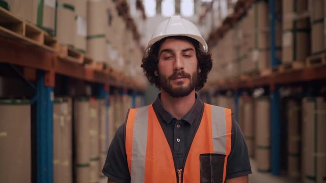 Portrait of a serious young warehouse worker looking at camera standing in between the rows of storage rack