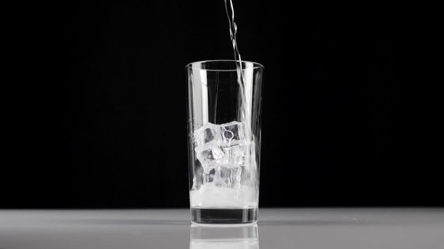 Pure fresh mineral water is poured into a transparent glass with ice cubes on a black background. Concept of stay at home and eat healthy food with soda and ice. Wide shot. Slow motion