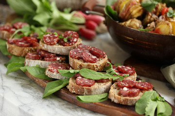 Summer snacks. Bread with sausage and herbs on a wooden board. A bunch of radishes, a pan with potatoes on a light table. Background image, copy space
