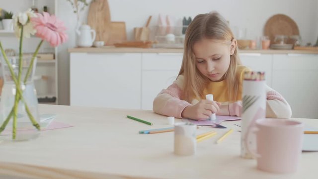 Lockdown of wonderful Caucasian girl with blond hair sitting at table and making greeting card for her mum using glue stick