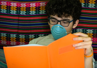 A bedridden Caucasian boy wears a blue protective mask. He is studying his orange-covered notebook....