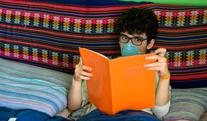 A bedridden Caucasian boy wears a blue protective mask. He is studying his orange-covered notebook....