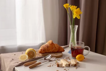 Poster In de kamer aan tafel waar het ontbijt wordt geserveerd met een kopje thee, croissants, citroenen en gele narcis in een glazen vaas © Natasha 