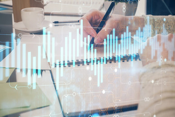 Hand taking notes in notepad. Forex chart holograms in front. Concept of research. Double exposure