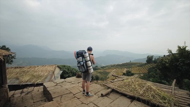 A young caucasian man tourist filming nepalese terraced rice fields from a hill. Backpack. Nepal, village.