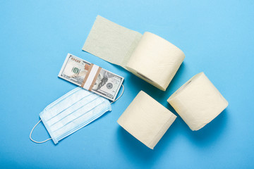 toilet paper rolls, medical mask and dollars banknotes on a blue background. Flat lay, top view. Concept hygiene product, virus, deficiency, quarantine