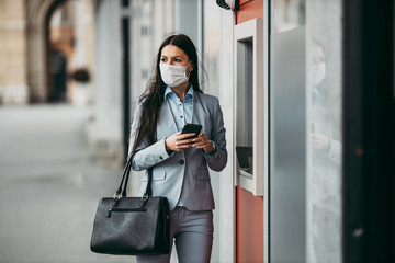 Elegant business woman with protective mask standing on city street and using ATM machine to...
