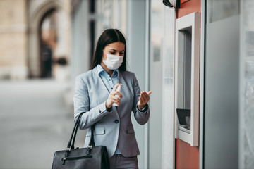 Elegant woman with protective mask standing on city street and using alcohol spray to disinfect her...
