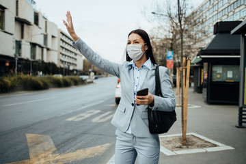Obraz premium Elegant business woman with protective mask standing alone on empty street and calling for bus or taxi transport with raised hand. Corona virus or Covid-19 lifestyle concept.