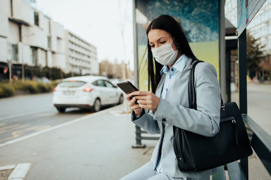 Elegant business woman with protective mask sitting alone on empty street and waiting for bus or taxi transport while using phone. Corona virus or Covid-19 lifestyle concept.