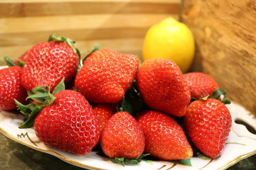 Close-up of a large ripe strawberry on a white and gold platter against a yellow lemon in the corner of a wooden wall. 