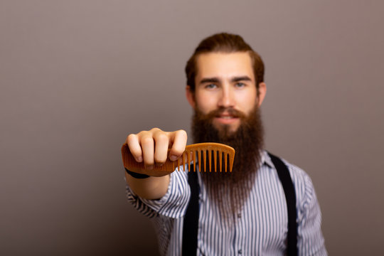 Man With A Long Beard On A Grey Background Holds A Comb, Portrait. Young Bearded Smiling Overgrown Man Holding Comb. Copy Space.