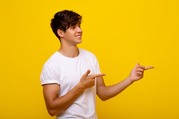 Smiling young man in casual white t-shirt posing isolated on bright yellow background. Studio portrait. Person sincere emotions, lifestyle concept. Copy space. Pointing index fingers aside.