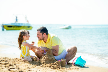 Fototapeta premium Dad and little girl play, sculpt sand castles on the beach and fool around on the sea coast. Family vacation at sea with a child.