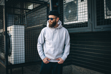 City portrait of handsome hipster guy with beard wearing gray blank hoodie or hoody and hat with...