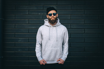 City portrait of handsome hipster guy with beard wearing gray blank hoodie or hoody and hat with...