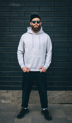 City portrait of handsome hipster guy with beard wearing gray blank hoodie or hoody and hat with space for your logo or design. Mockup for print