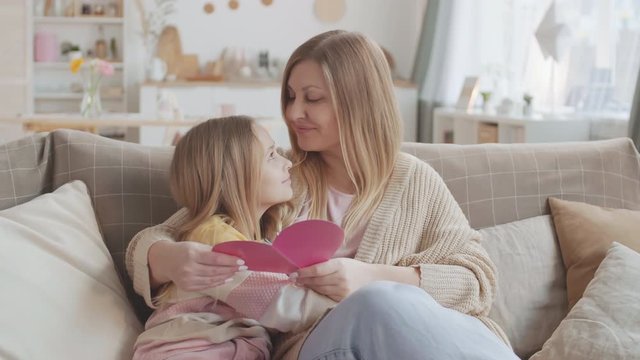 Medium shot of mother greeting her little daughter for handmade heart-shaped card by hugging and kissing her