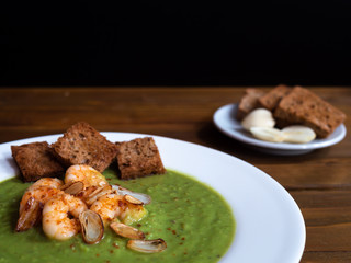 A plate of thick fresh green pea soup with prawns on a rustic wooden table with a garlic and croutons on the side. Close up.