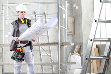 woman construction worker builder looking at bluprint, wearing helmet, hearing protection headphones and tools belt bag in building site indoors background