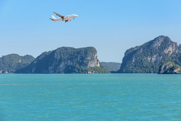 Fototapeta na wymiar White passenger airplane landing above tropical blue sea over small limestone island with blue sky travel destinations concept