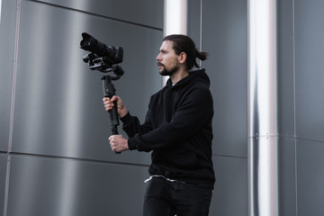 Young Professional videographer holding professional camera on 3-axis gimbal stabilizer. Pro equipment helps to make high quality video without shaking. Cameraman wearing white hoodie making a videos. © Volodymyr_sh