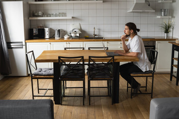Freelancer working from home on the kitchen and using laptop. Bearded man working with a laptop and reading good news. Handsome successful self entrepreneur sitting and working at his modern home.