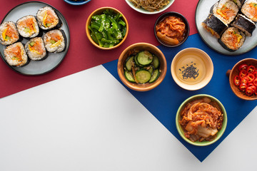 top view of plates with fresh gimbap near korean side dishes on white, blue and crimson