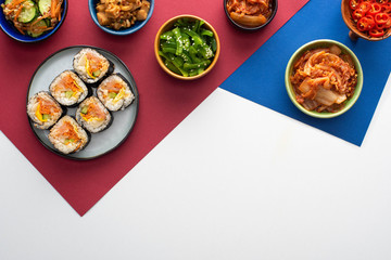 top view of plate with tasty gimbap near korean side dishes on white, blue and crimson