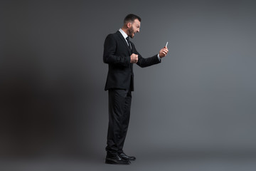 profile of angry businessman in suit screaming while looking at smartphone on grey