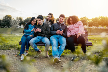 Group of young friends at the park playing with smart phones sitting on a bench at sunset -...